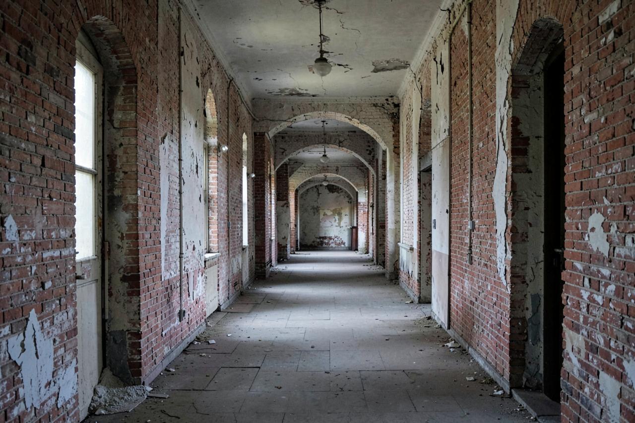 A derelict brick corridor with arched doorways, peeling plaster and dusty light